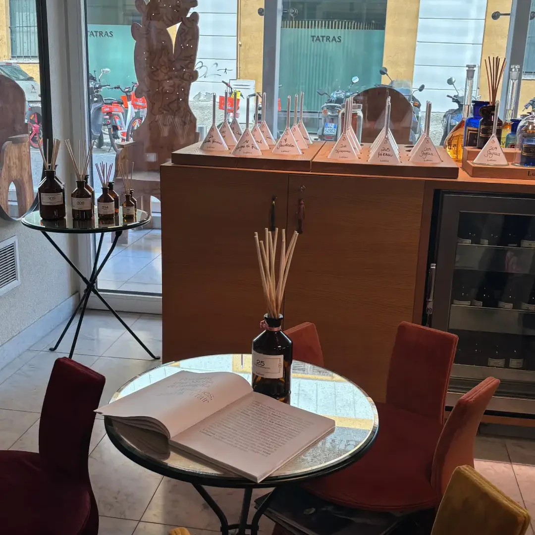 interno dello store di profumi con un tavolino con appoggiato un libro aperto, una cassettiera in legno, una sedia e un tavolo con appoggiati dei profumatori ambiente