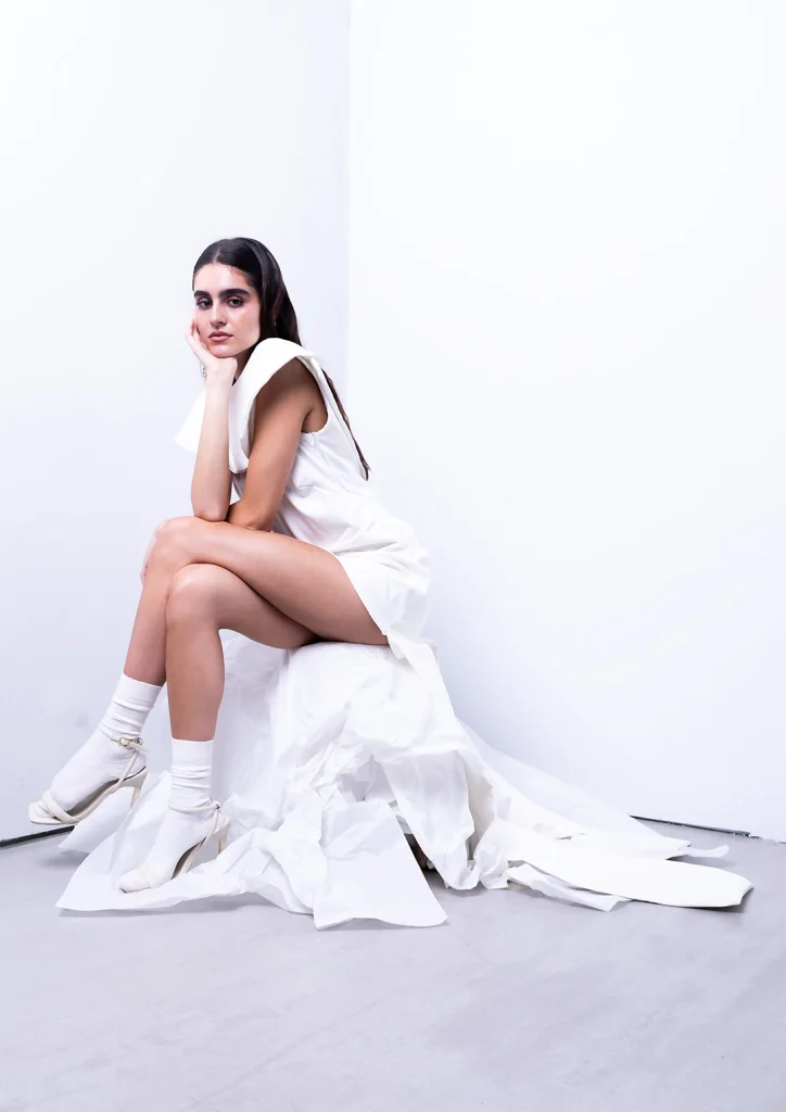 Model posing seated in the studio wearing a white dress with voluminous drapes.