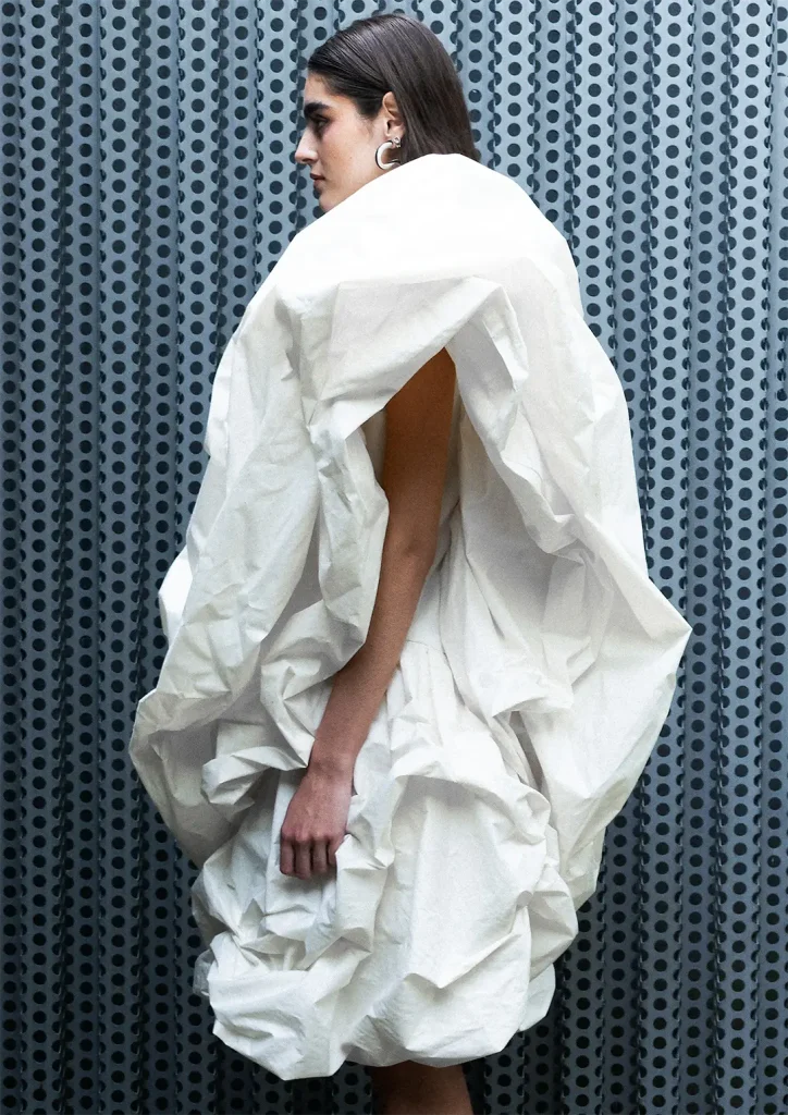 Model posing in an industrial setting wearing a white dress with voluminous drapes.